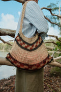 A person outdoors wearing a large woven shoulder bag with a natural straw base and dark brown and rust-red geometric patterned bands, photographed from behind against branches and water.