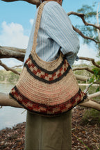 A person outdoors wearing a large woven shoulder bag with a natural straw base and dark brown and rust-red geometric patterned bands, photographed from behind against branches and water.