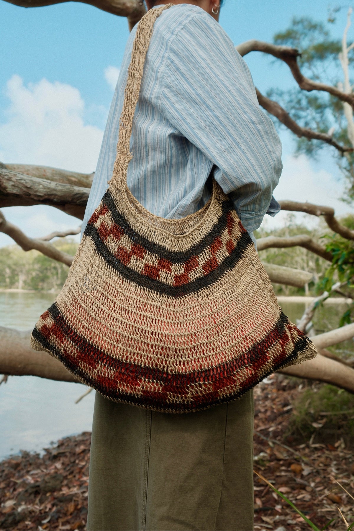 A person outdoors wearing a large woven shoulder bag with a natural straw base and dark brown and rust-red geometric patterned bands, photographed from behind against branches and water.