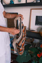 A person in a white shirt holds up a chunky knit shoulder bag with a geometric pattern in beige, orange, and black, set against a home decor background.
