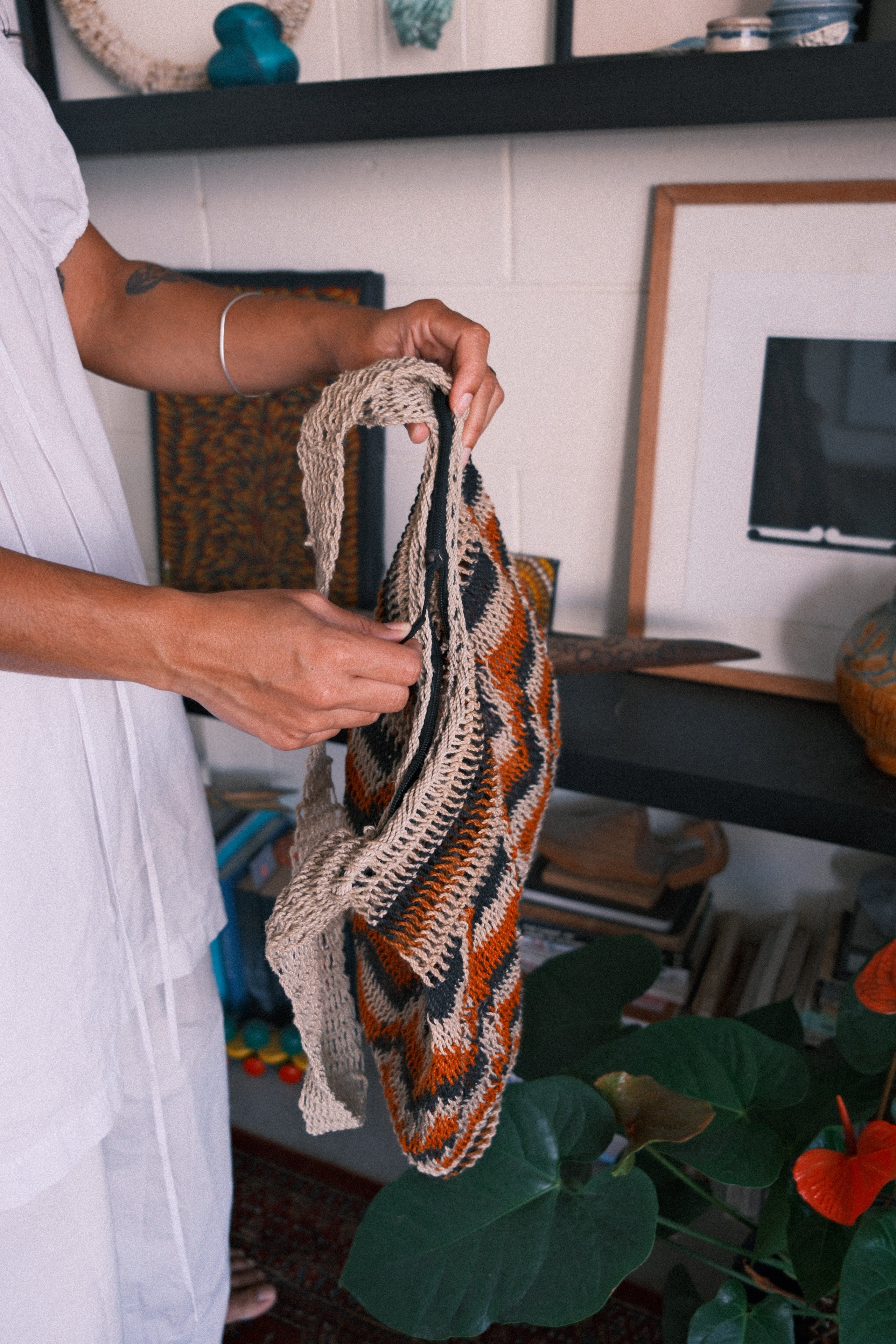 A person in a white shirt holds up a chunky knit shoulder bag with a geometric pattern in beige, orange, and black, set against a home decor background.